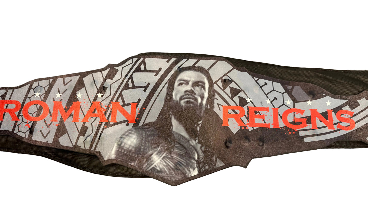 A custom WWE-style championship belt featuring Roman Reigns, laid flat on a textured stone floor. The belt has a black base with a large graphic of Roman Reigns in grayscale, red text reading 'ROMAN REIGNS', and white geometric patterns.
