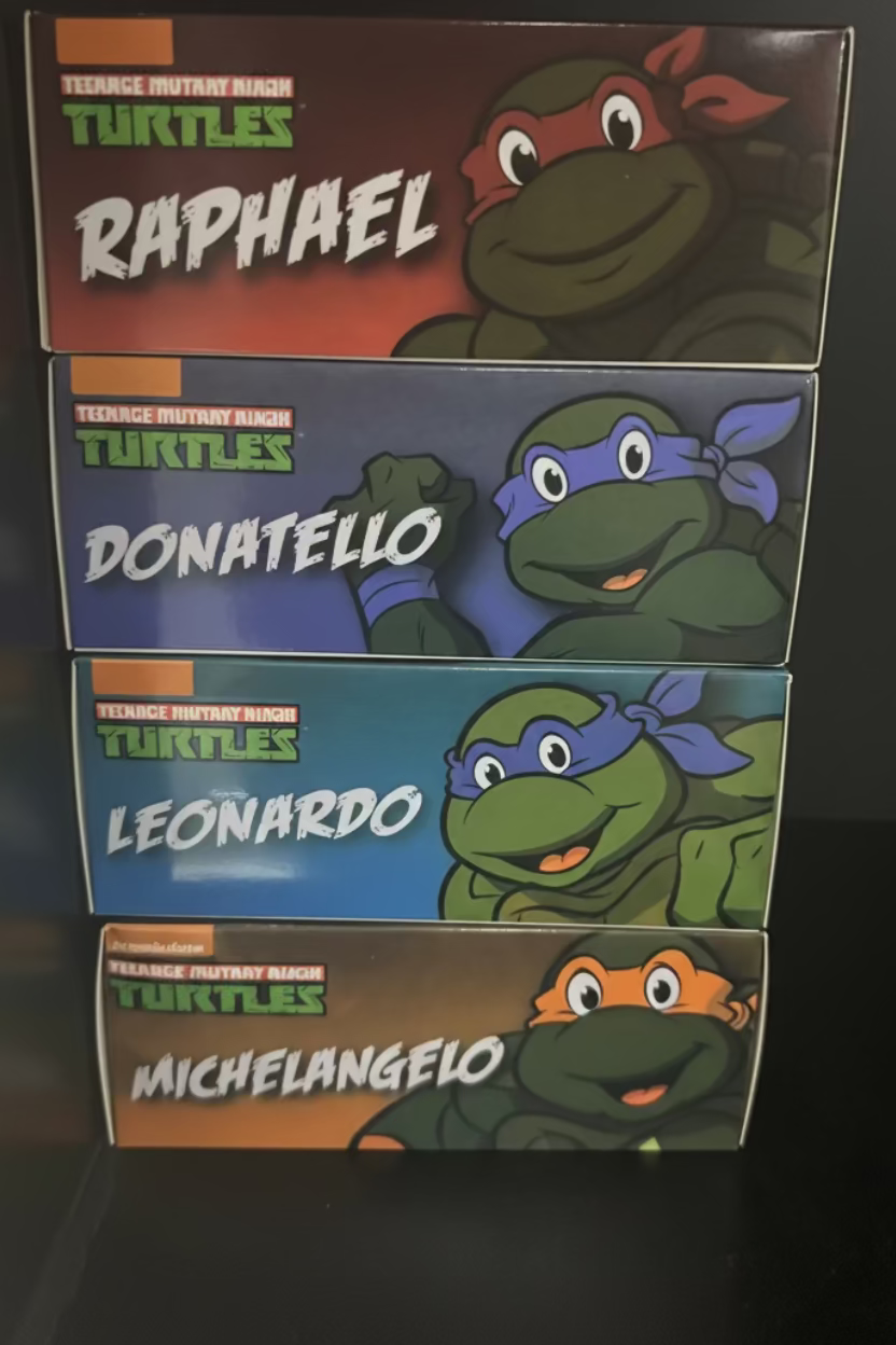 Four Teenage Mutant Ninja Turtles character boxes stacked vertically on a dark surface. The boxes feature Raphael, Donatello, Leonardo, and Michelangelo, each with their own colored background and character illustration.