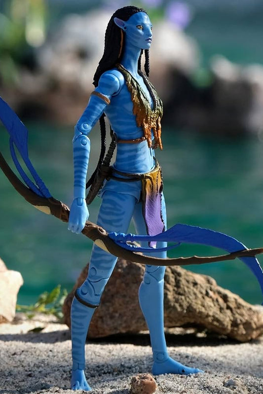 A blue humanoid alien action figure standing on a sandy shore with rocks and turquoise water in the background, holding a curved brown bow with bright blue strings.