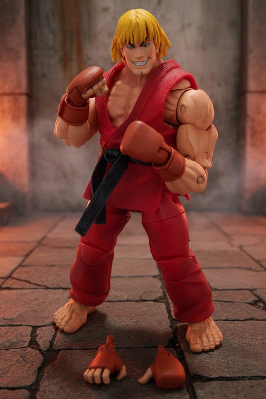 A Street Fighter character action figure of Ken Paret in a fighting pose, wearing a red gi with a black belt and brown gloves, standing on a white surface against a white background with an extra pair of hands placed in front.